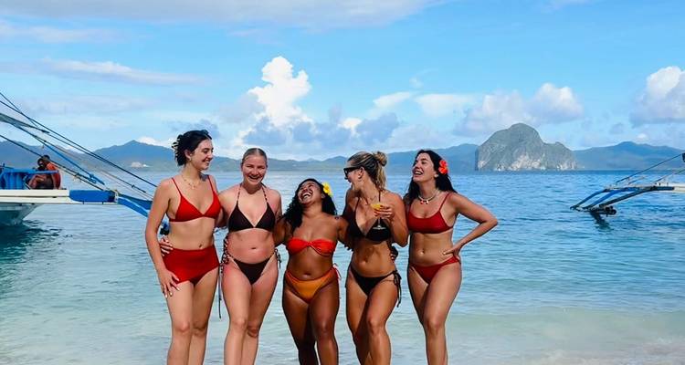 Fünf Freunde in leuchtend farbigen Bikinis, die an einem Strand mit weißem Sand lachen, mit blauen Auslegerkanus und Inseln im Hintergrund.