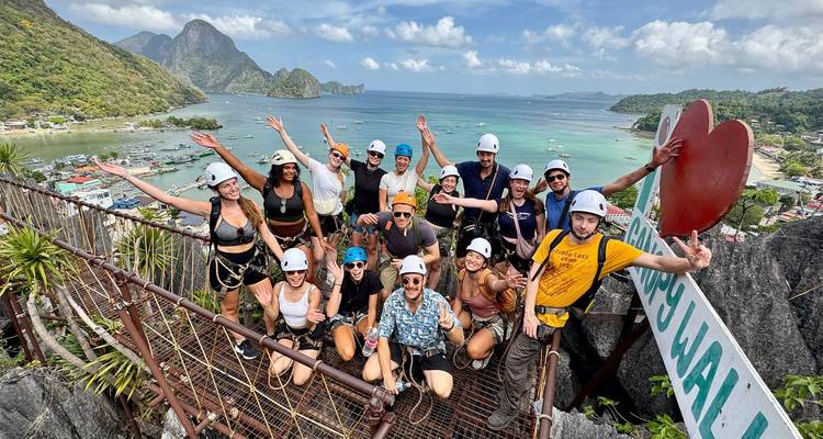 Große Abenteurergruppe mit Helmen und Sicherheitsgeschirren, die auf einer Aussichtsplattform auf einem Klippenrand jubelt und turquisfarbene Buchten sowie Kalksteininseln überblickt.