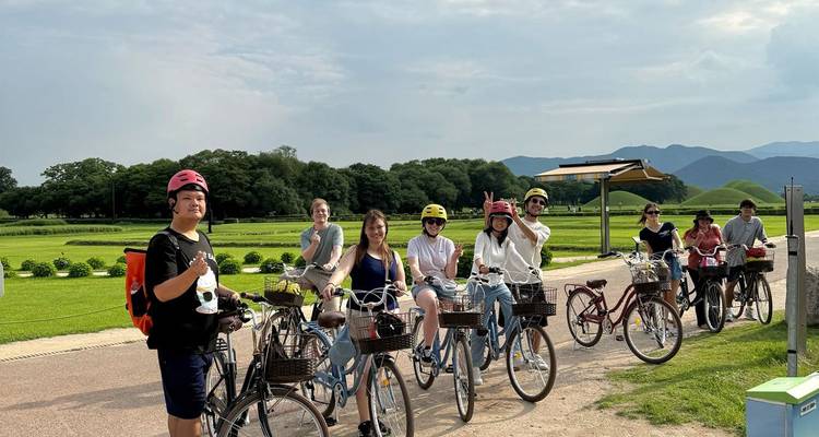Cyclists pause on their bikes along a path in a grassy park dotted with ancient burial mounds.