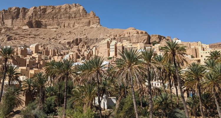 Oasis de palmeras verdoso ubicado frente a casas de adobe apiladas y acantilados desérticos escarpados bajo un cielo despejado.