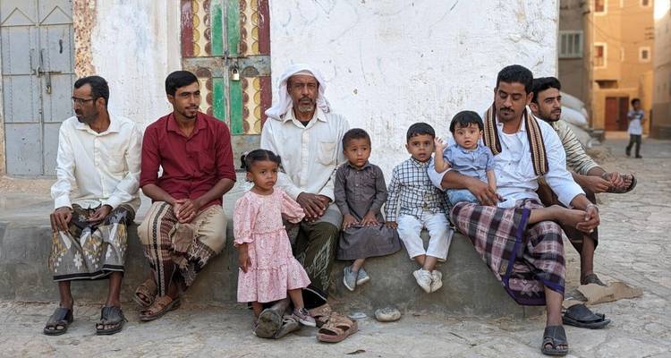 Hombres locales y niños con vestimenta tradicional se sientan socializando contra una pared encalada.