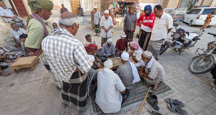 Hombres ancianos se reúnen alrededor de un juego de mesa en una calle polvorienta del pueblo, observados por espectadores.