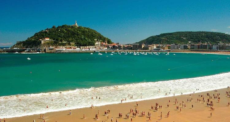 Une scène de plage animée avec des gens sur le sable et une baie turquoise, encadrée par des collines luxuriantes en arrière-plan.