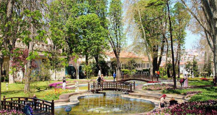 Des gens se promènent dans un jardin avec des fontaines et des arbres par une journée ensoleillée.