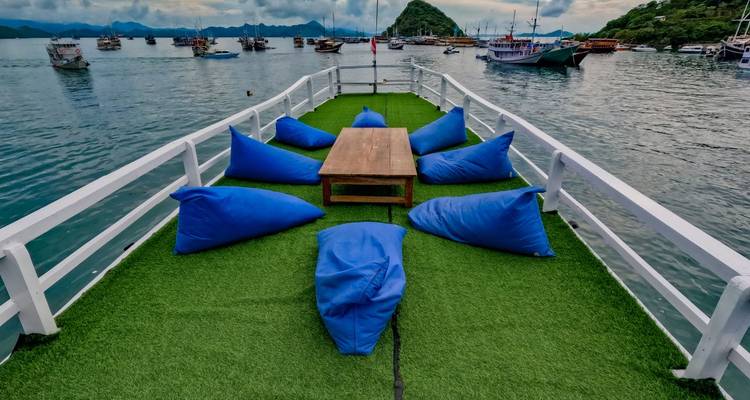 Pont ouvert d'un bateau aménagé avec du gazon artificiel vert, des poufs bleus et une table en bois donnant sur un port avec des bateaux amarrés et des îles.