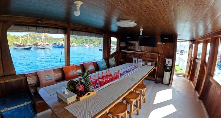 Salon à manger lambrissé de bois sur un bateau avec une longue table commune, des coussins colorés et des fenêtres ouvrant sur la vue mer.