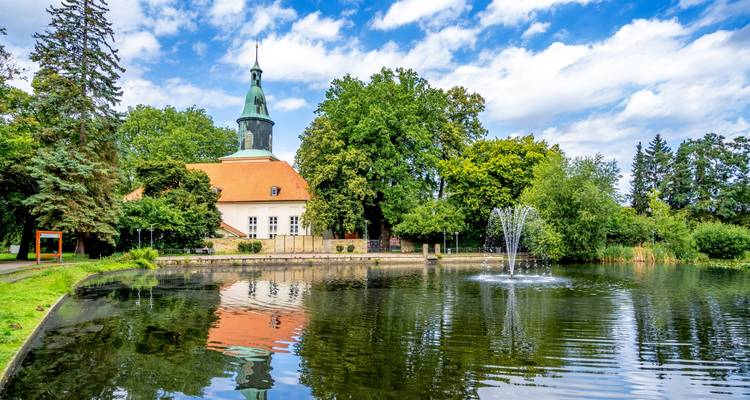 Parc pittoresque avec une église, une fontaine et un bassin de réflexion sous un ciel bleu.