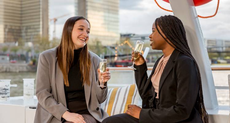 Twee vrouwen die genieten van drankjes op een cruise met stadsgebouwen op de achtergrond.