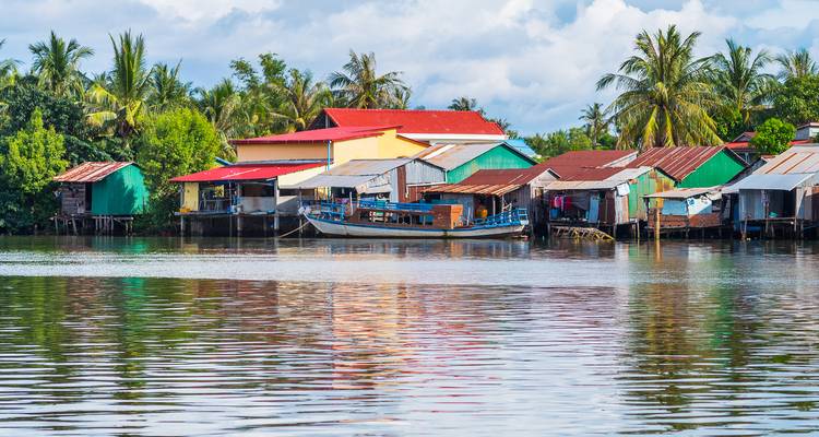 Casas coloridas sobre pilotes junto al río y botes se reflejan en aguas tranquilas bordeadas por palmeras.