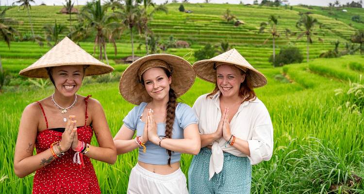 Drie jonge vrouwen in traditionele hoeden poseren met hun handen tegen elkaar in een zonnig rijstveld