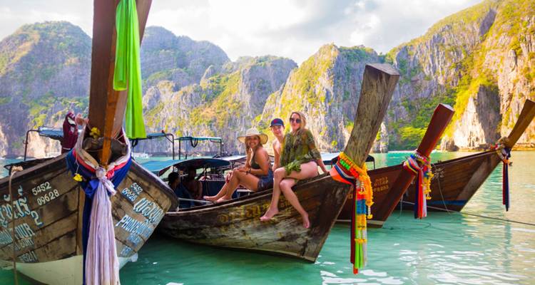 Travellers sit on decorated long-tail boats in a jade green bay surrounded by limestone cliffs