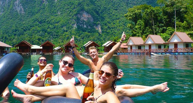 Grupo de jóvenes viajeros relajándose en flotadores con bebidas en un lago rodeado de colinas cubiertas de selva y cabañas flotantes.