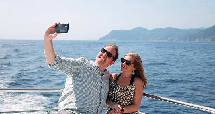 Couple prenant un selfie sur un bateau avec la mer et les collines en arrière-plan.
