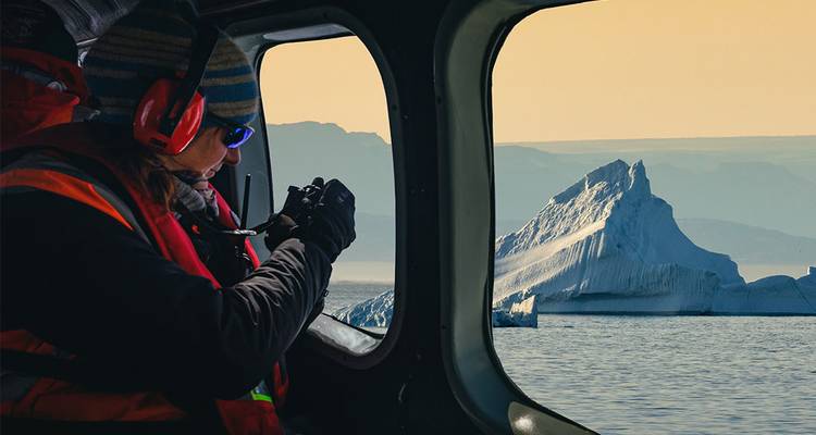 A passenger wearing headphones photographs a towering iceberg through the window of a helicopter at golden hour.