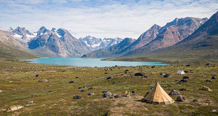 Tents are pitched beside a turquoise alpine lake surrounded by jagged snow-dusted peaks in Greenland’s wilderness.