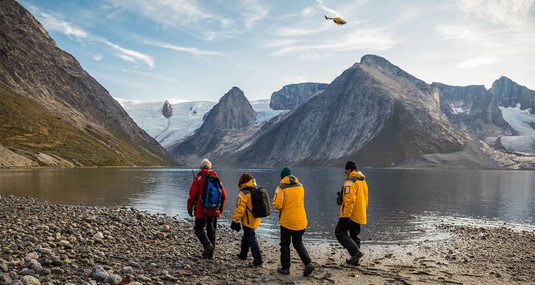Travellers in yellow jackets walk along a glacier-fed lake shore with steep granite walls while a helicopter hovers above.