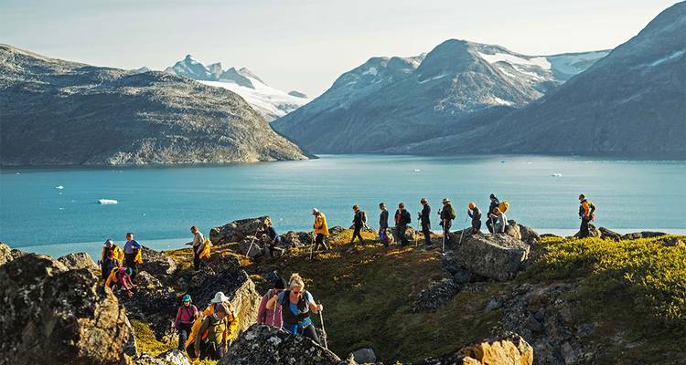 Un grand groupe de randonneurs traverse un terrain rocheux surplombant un fjord bleu encadré par des montagnes aux sommets enneigés.