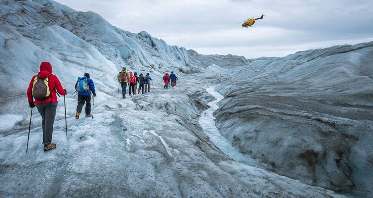 Des voyageurs portant des crampons marchent en file indienne à travers un glacier bleu crevassé tandis qu'un hélicoptère vole au-dessus.