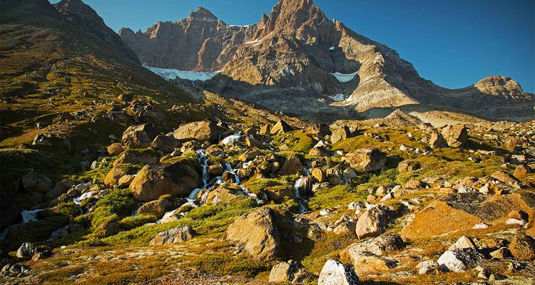 Une lumière dorée baigne un paysage alpin accidenté de rochers, de prairies herbeuses et de pics déchiquetés avec un glacier au loin.