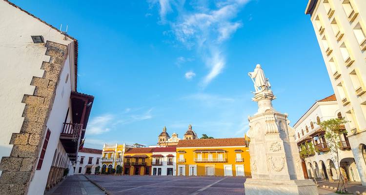 Bright colonial plaza with statue, colorful facades and cathedral domes under a vivid blue Cartagena sky.