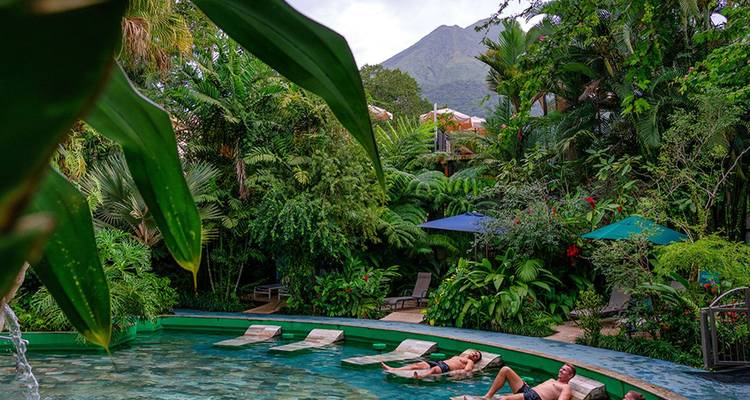Los viajeros se relajan en una piscina termal exuberante con el Volcán Arenal elevándose más allá de las copas de los árboles.