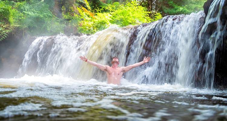 Un hombre levanta alegremente los brazos mientras se baña bajo una pequeña y exuberante cascada de la selva tropical.