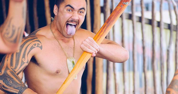 Artista maorí con tatuajes tradicionales saca la lengua mientras sostiene un remo de madera.