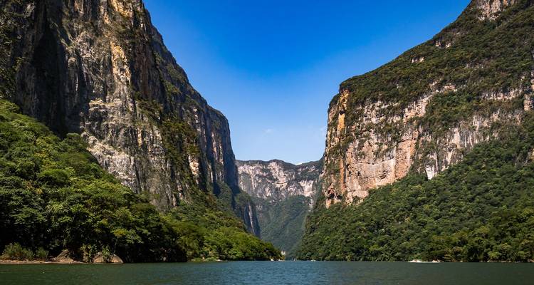 Steile bosrijke canyonwanden die hoog boven een rustige groene rivier uitsteken onder een helderblauwe lucht.