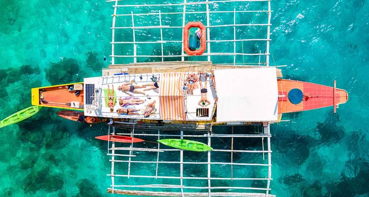 Vue aérienne prise par drone d'un bateau à balancier traditionnel philippin avec des baigneurs et des kayaks dans une eau turquoise éclatante.