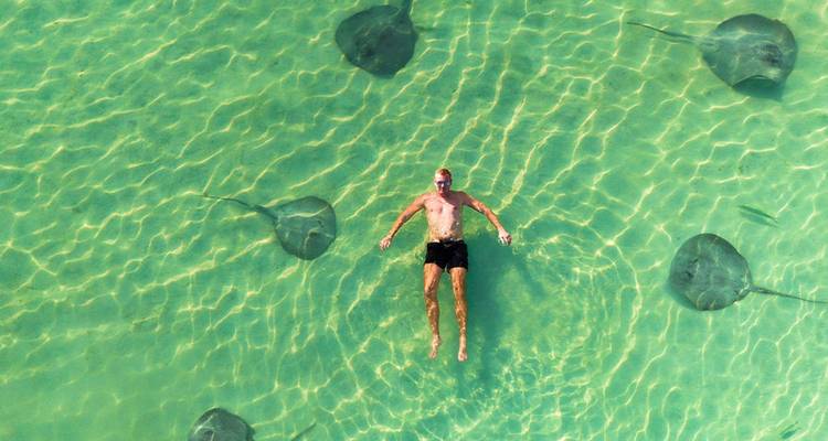 Un hombre flota en aguas cristalinas de color turquesa rodeado de graciosas rayas vistas desde arriba.