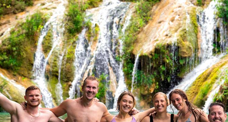 Amigos riendo metidos hasta la cintura en una piscina de la selva con cascadas de varios niveles detrás.
