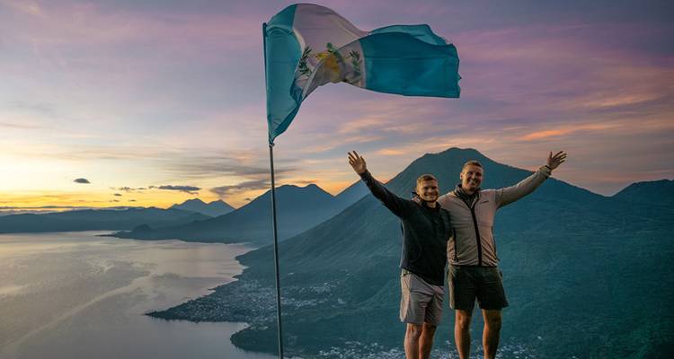 Dos excursionistas agitan una bandera guatemalteca sobre el Lago Atitlán en un amanecer colorido.