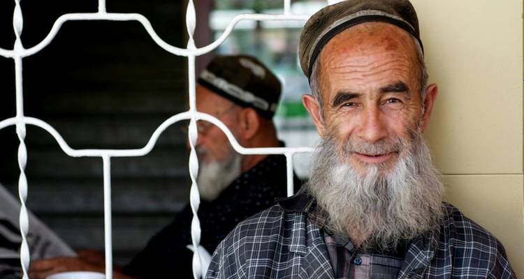 Portrait of an elderly man with traditional headwear smiling, another person reading a newspaper in the background.