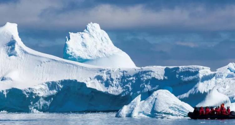Una pequeña barca inflable con pasajeros bien abrigados flota junto a enormes témpanos de hielo azul y blanco bajo un cielo parcialmente nublado.