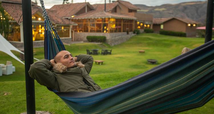 Man relaxes in a striped hammock on a grassy lodge lawn at dusk with mountains beyond.