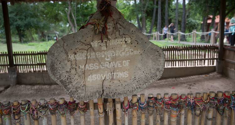 Letrero hecho de una sección de tronco de árbol que señala un sitio de fosa común, rodeado de una cerca de bambú y ofrendas de pulseras.