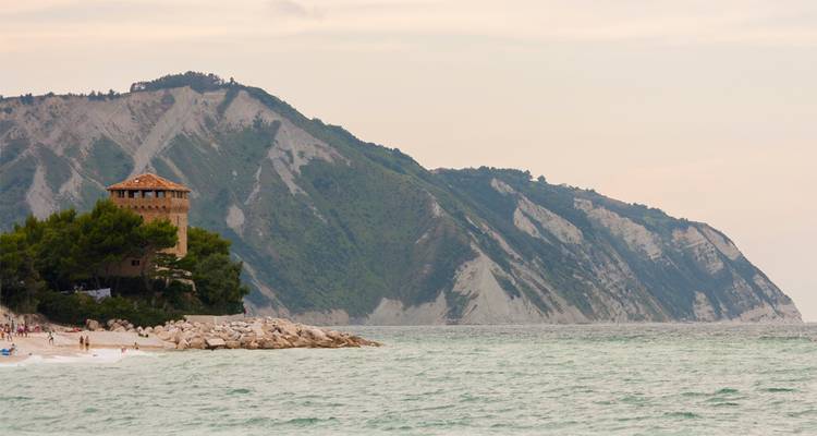 Stone watchtower on forested promontory with steep cliffs rising above a tranquil sea.