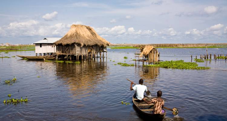 Canoe with two people paddling through village on stilts over water.
