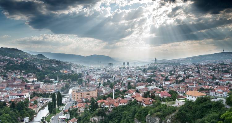 Luchtfoto van Sarajevo in een groene vallei met zonnestralen die door dramatische wolken boven de daken van de stad breken.