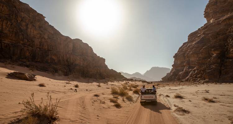 Open-back Toyota jeep carrying travellers along a sandy track between towering desert cliffs with the sun high above.