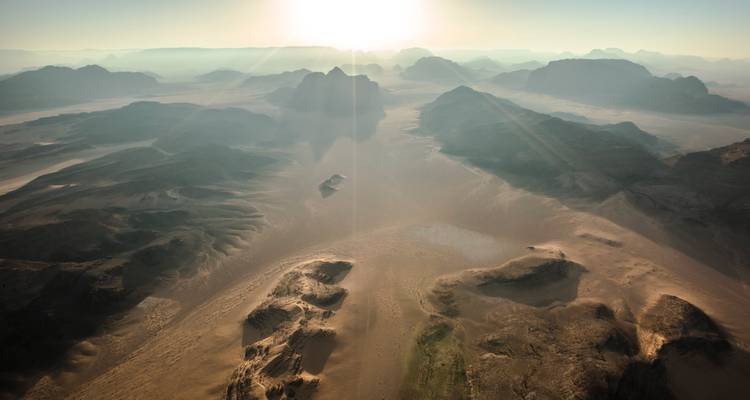 Sweeping aerial view of sun-lit desert mountains and valleys at dawn with soft haze over Wadi Rum.
