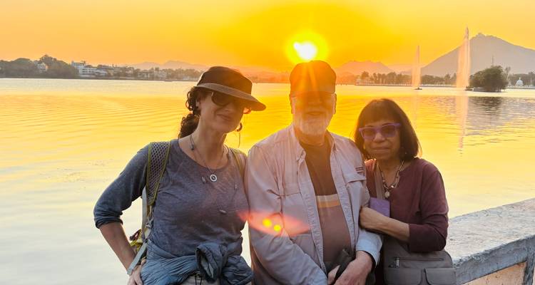 Drei Reisende posieren auf einer Seepromenade mit Springbrunnen, ruhigem Wasser und fernen Hügeln, die von einem leuchtend orangen Sonnenuntergang beleuchtet werden.