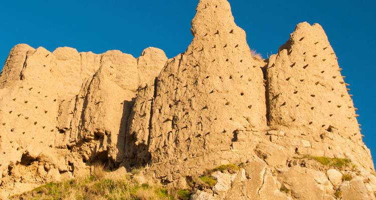 Ruinas de una fortaleza de tierra bajo un sol radiante, con muros dentados y pequeños agujeros para nidos, recortadas contra un cielo azul vibrante.