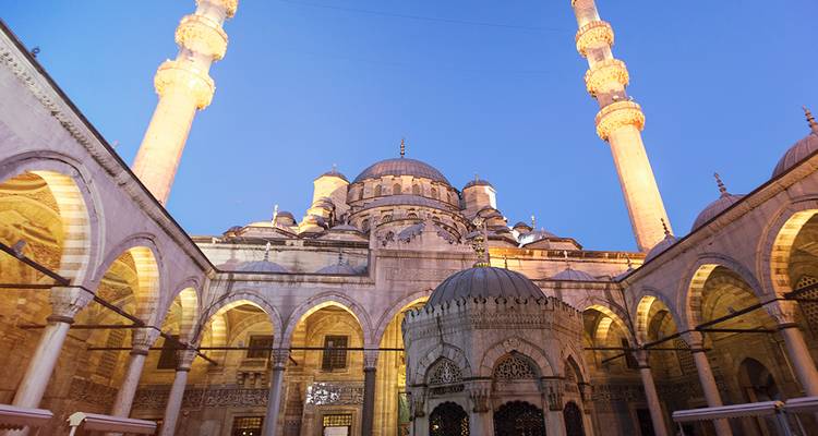 Patio de una mezquita otomana iluminada con arcos elegantes y dos minaretes brillando contra un cielo azul profundo al atardecer.