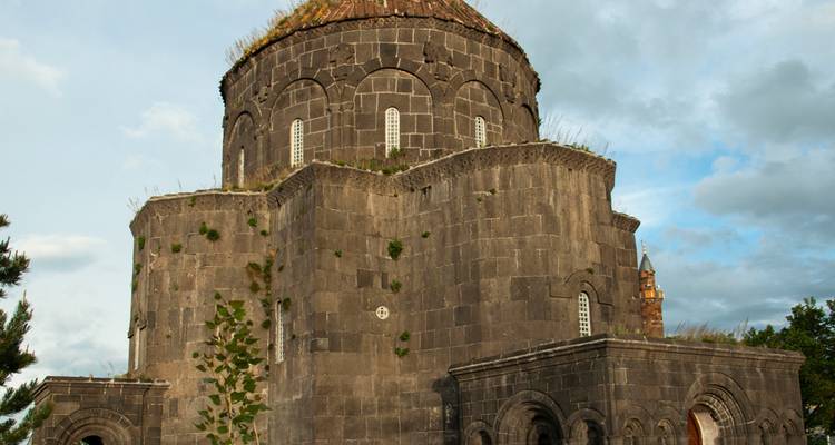 Iglesia medieval redonda de piedra con ventanas arqueadas y parches de vegetación en el techo, ubicada bajo un cielo parcialmente nublado.