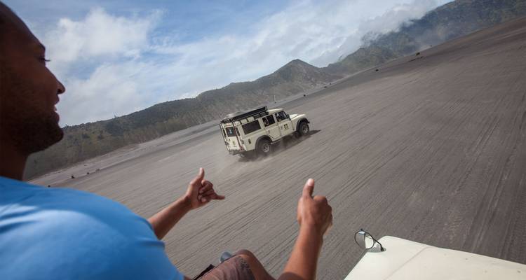 Un viajero levantando el pulgar desde una jeep mientras otro vehículo acelera a través de una llanura de arena volcánica con montañas de fondo.