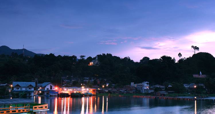 Vista del atardecer con las luces del pueblo reflejadas en un lago tranquilo rodeado de colinas boscosas.