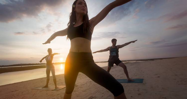 Un petit groupe pratiquant des postures de yoga sur une plage de sable au coucher du soleil avec l'océan en arrière-plan.