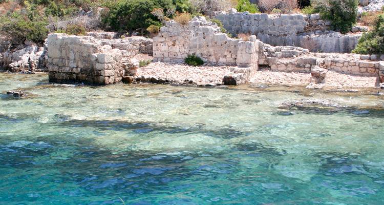 Crystal-clear turquoise water laps against submerged stone ruins along the rocky coast of Kekova.