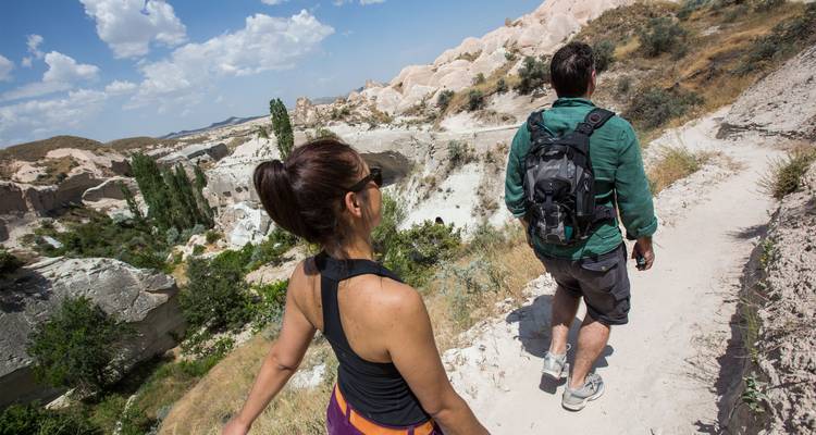 Two hikers walking along a dusty trail between pale rock formations and sparse vegetation under a bright sky.